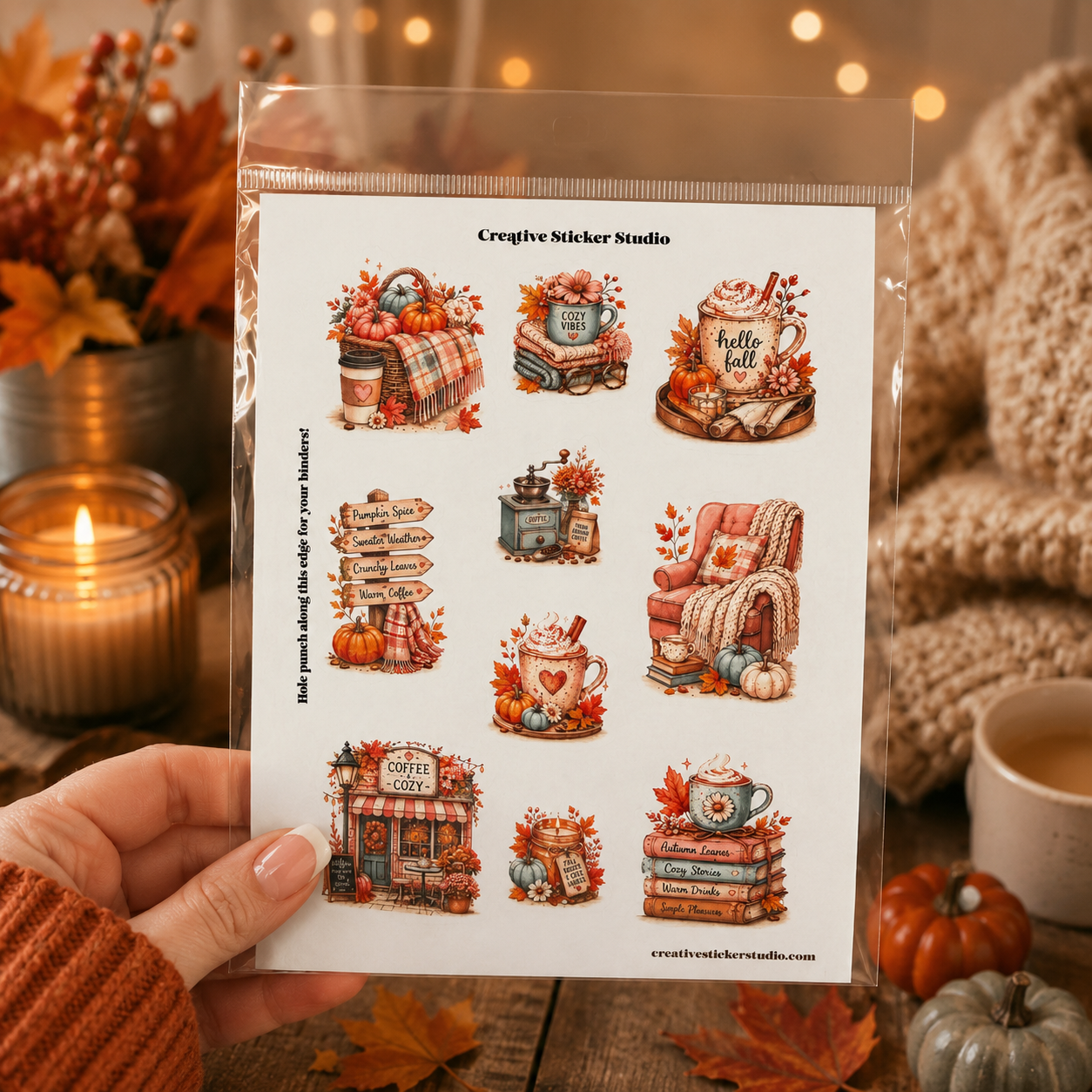A hand holds a clear plastic sleeve containing a sheet of nine autumn-themed coffee and tea stickers. The background is a cozy, warm fall scene with a lit candle, dried leaves, a mug, and a knitted sweater.