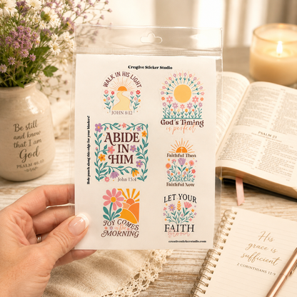 A hand holds a clear plastic package containing six colorful Christian-themed sticker designs with Bible verses and floral sun motifs. The scene is styled with an open book, a candle, a ceramic vase with flowers, and a notebook in the background.
