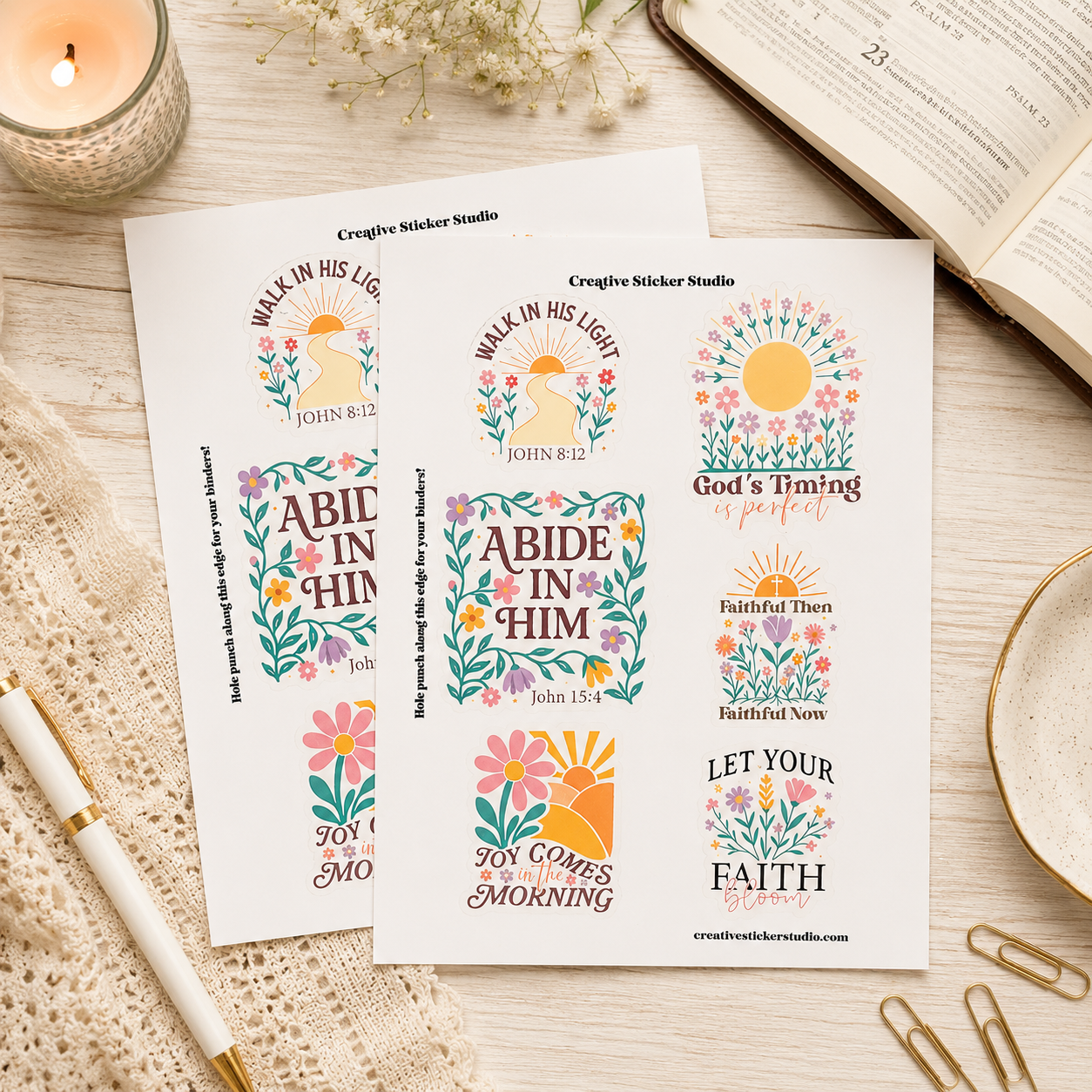 Two sticker sheets with colorful Bible verse designs and floral sun motifs, styled on a light wood surface with an open book, candle, pen, paper clips, and dried flowers.
