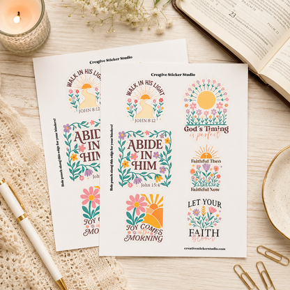 Two sticker sheets with colorful Bible verse designs and floral sun motifs, styled on a light wood surface with an open book, candle, pen, paper clips, and dried flowers.