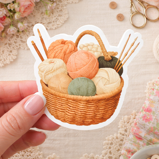 Sticker of a woven basket with yarn balls and knitting needles held by a hand, with a floral and textured background.