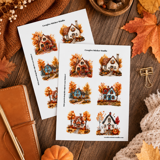 Two printed sticker sheets featuring watercolor-style autumn houses, styled on a wooden surface with fall leaves, a knit blanket, a notebook, and small decorative props.