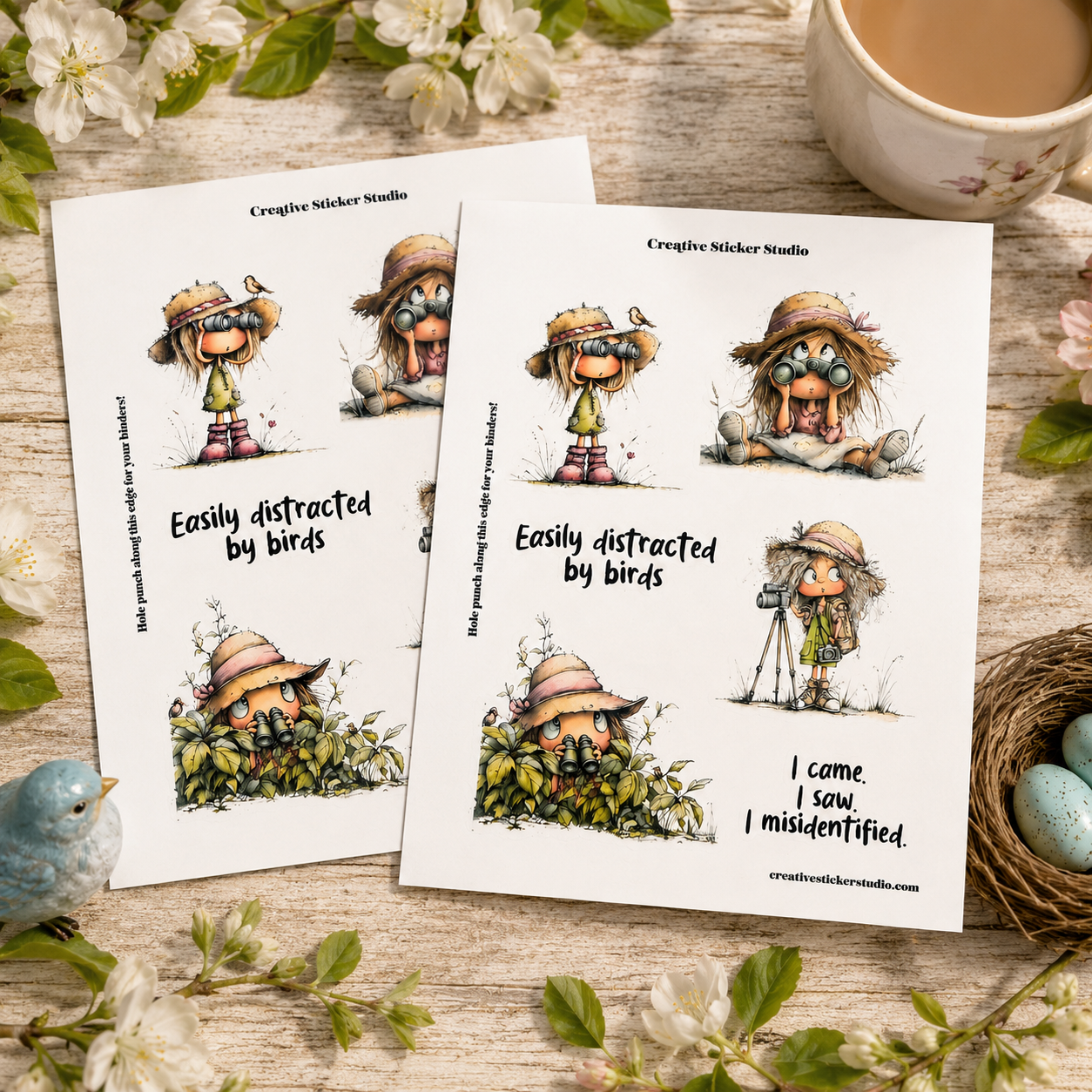 Two printed sticker sheets with illustrated cartoon characters wearing straw hats and the text 'Easily distracted by birds' and 'I came, I saw, I missed identified.' are arranged on a wooden surface with flowers, a cup of tea, a small bird, and a nest with blue eggs around them.