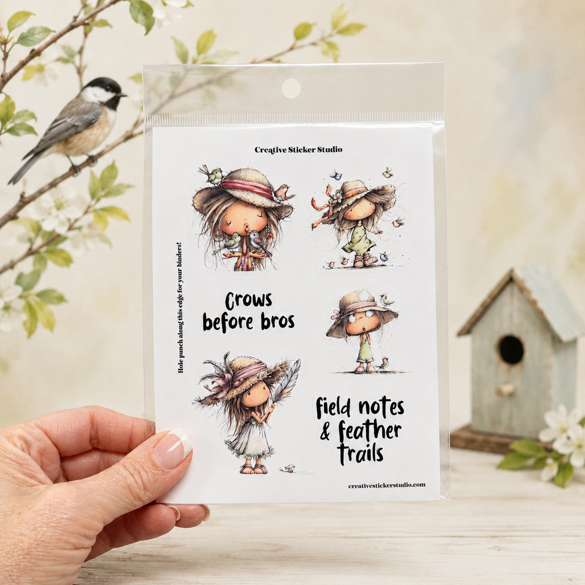 A hand holds a clear plastic package of Creative Sticker Studio 'Crows before bros' clear stamps, featuring four illustrated cartoon crows. The background is a soft-focus, warm-toned setting with a bird, a small birdhouse, and white blossoms.