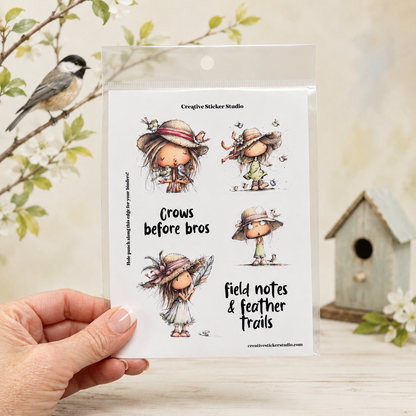 A hand holds a clear plastic package of Creative Sticker Studio 'Crows before bros' clear stamps, featuring four illustrated cartoon crows. The background is a soft-focus, warm-toned setting with a bird, a small birdhouse, and white blossoms.