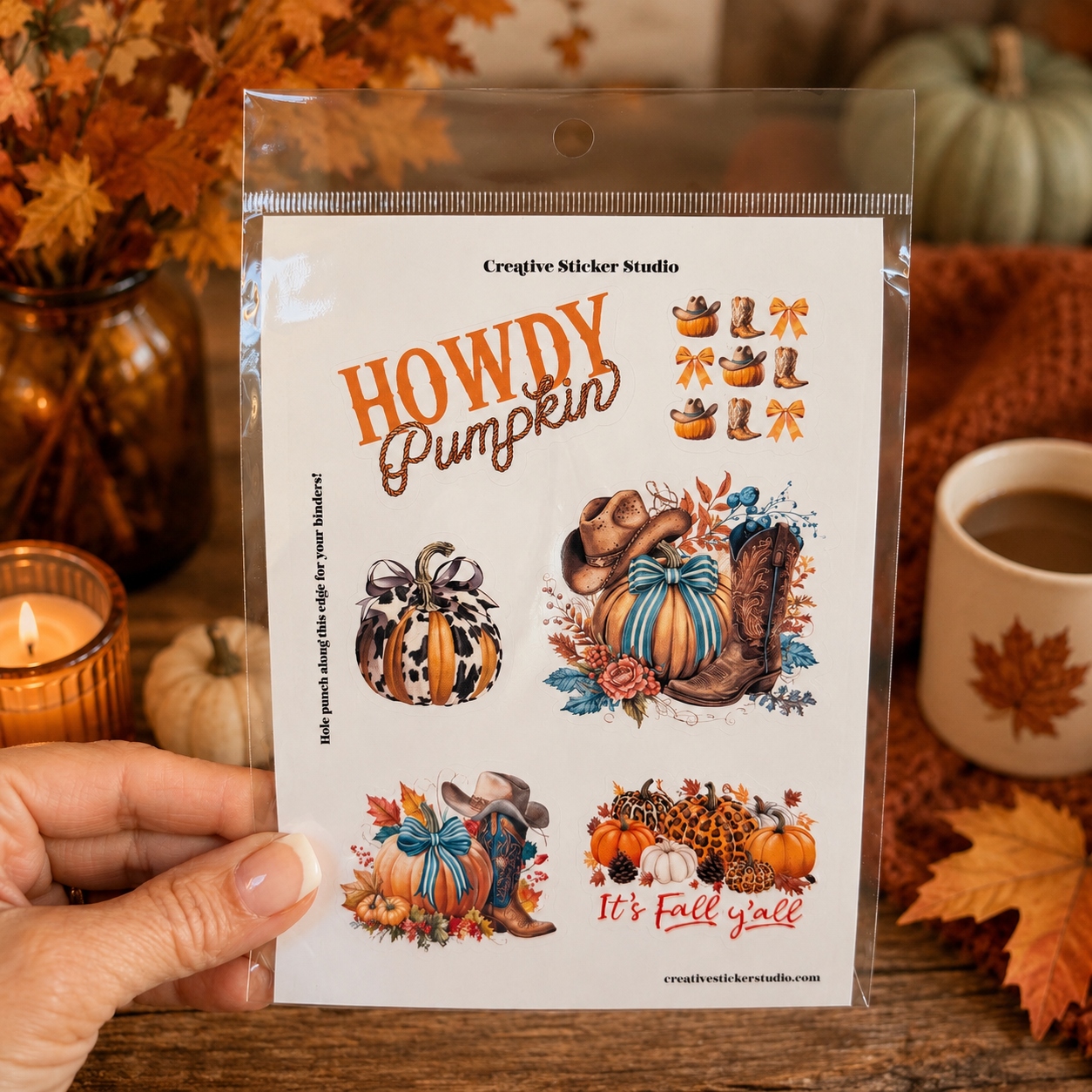 A hand holds a clear plastic package containing four Creative Sticker Studio 'Howdy Pumpkin' autumn-themed stickers, set against a cozy fall background with pumpkins, a candle, and a mug of coffee.