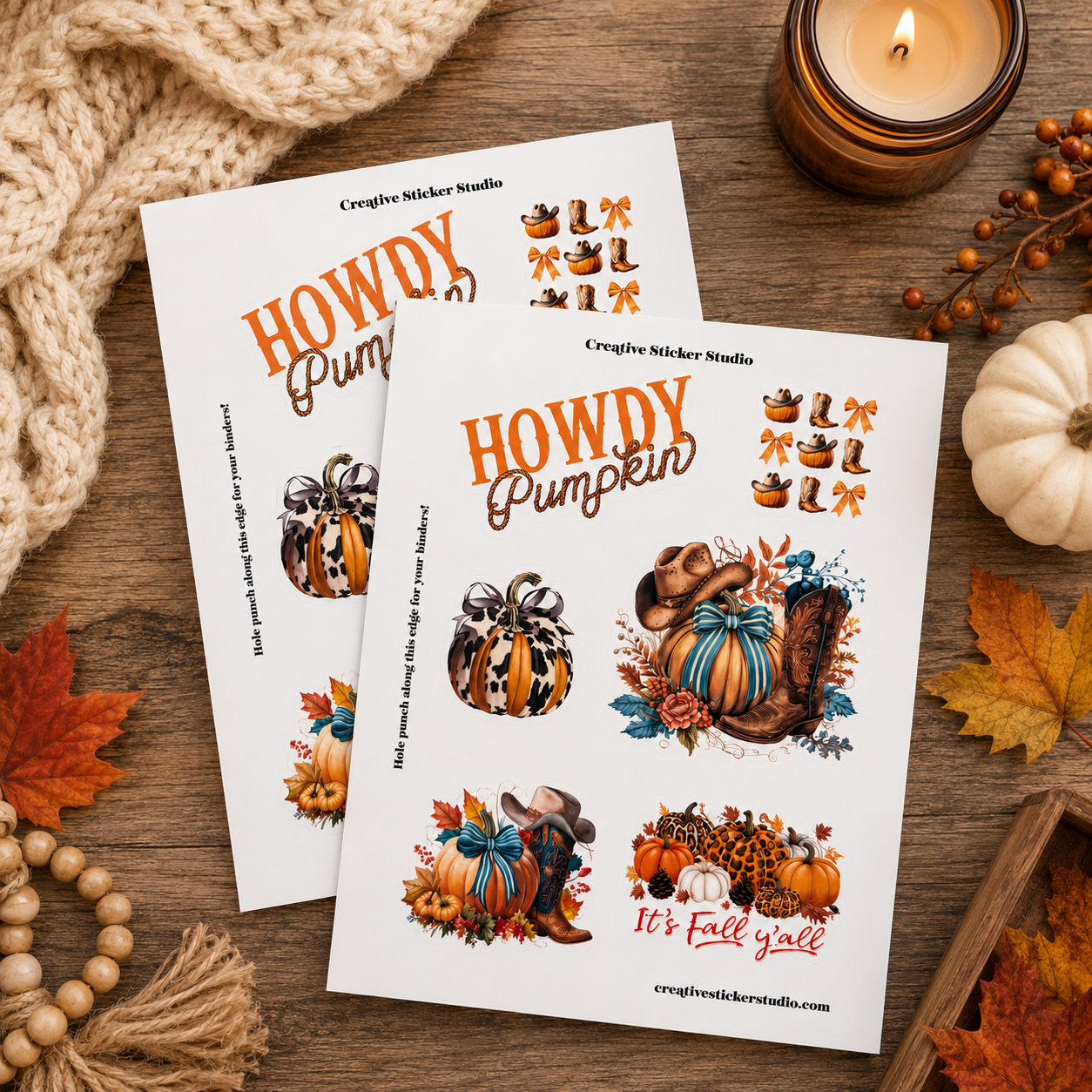Two printed sticker sheets with autumn-themed designs titled 'Howdy Pumpkin' are arranged on a wooden surface with a knitted blanket, candle, small pumpkin, fall leaves, and beads around them. The sheets show illustrated pumpkins, acorns, leaves, and fall motifs in orange, brown, blue, and cream tones.