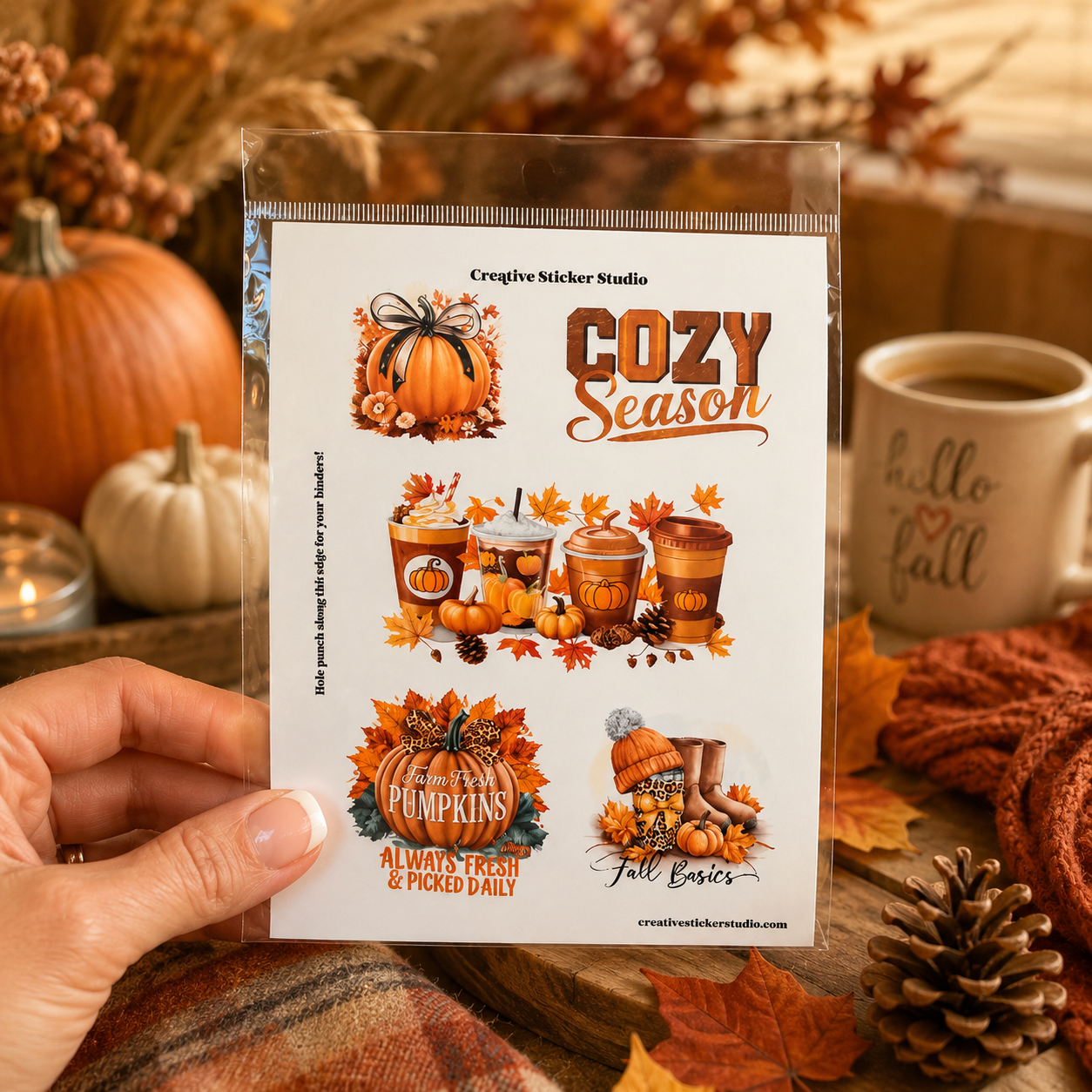 A hand holds a clear plastic sleeve containing a sheet of autumn-themed sticker designs with the text 'Cozy Season' and fall motifs like pumpkins, leaves, coffee cups, and boots. The background is a cozy, warm autumn scene with pumpkins, a mug that says 'hello fall,' and a scarf.
