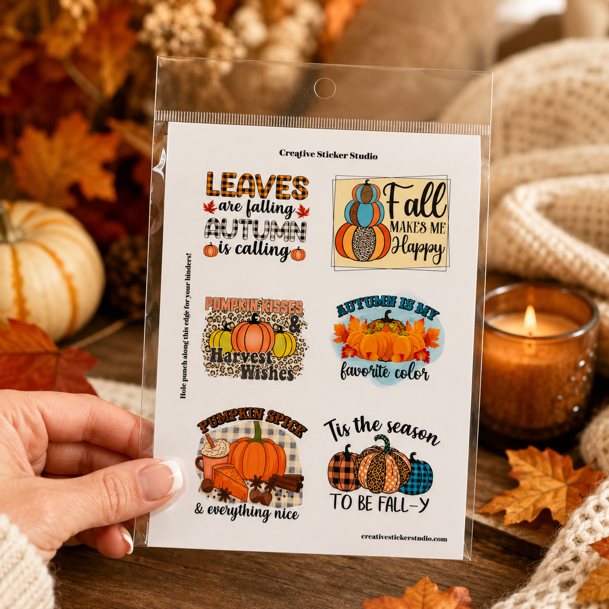 A hand holds a clear plastic package containing a sheet of Creighton Studio sticker designs with autumn-themed quotes and pumpkin illustrations, set against a cozy fall background with leaves, a candle, and knitted fabric.