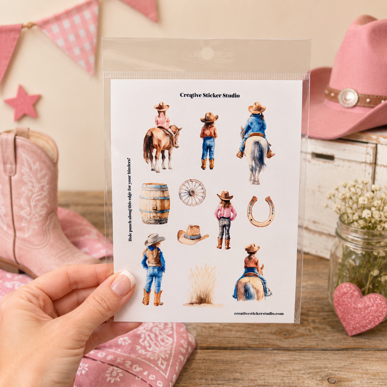 A hand holds a clear plastic bag containing a sheet of western-themed sticker designs, including cowboy and cowgirl figures, a barrel, wagon wheel, horseshoe, and hats. The background is styled with pink cowboy boots, a pink hat, bunting, and a rustic wooden surface.