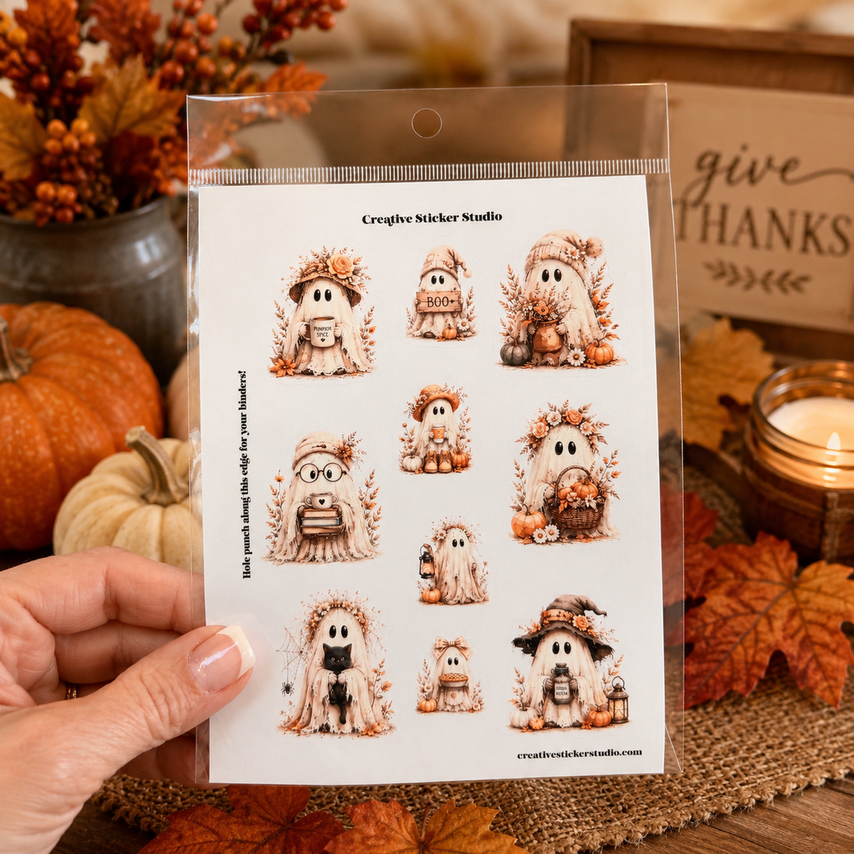 A hand holds a clear plastic sheet containing nine small Halloween-themed ghost illustrations with autumn decorations. The sheet is labeled 'Creative Sticker Studio' and is photographed in a fall-themed setting with pumpkins, leaves, and a candle in the background.