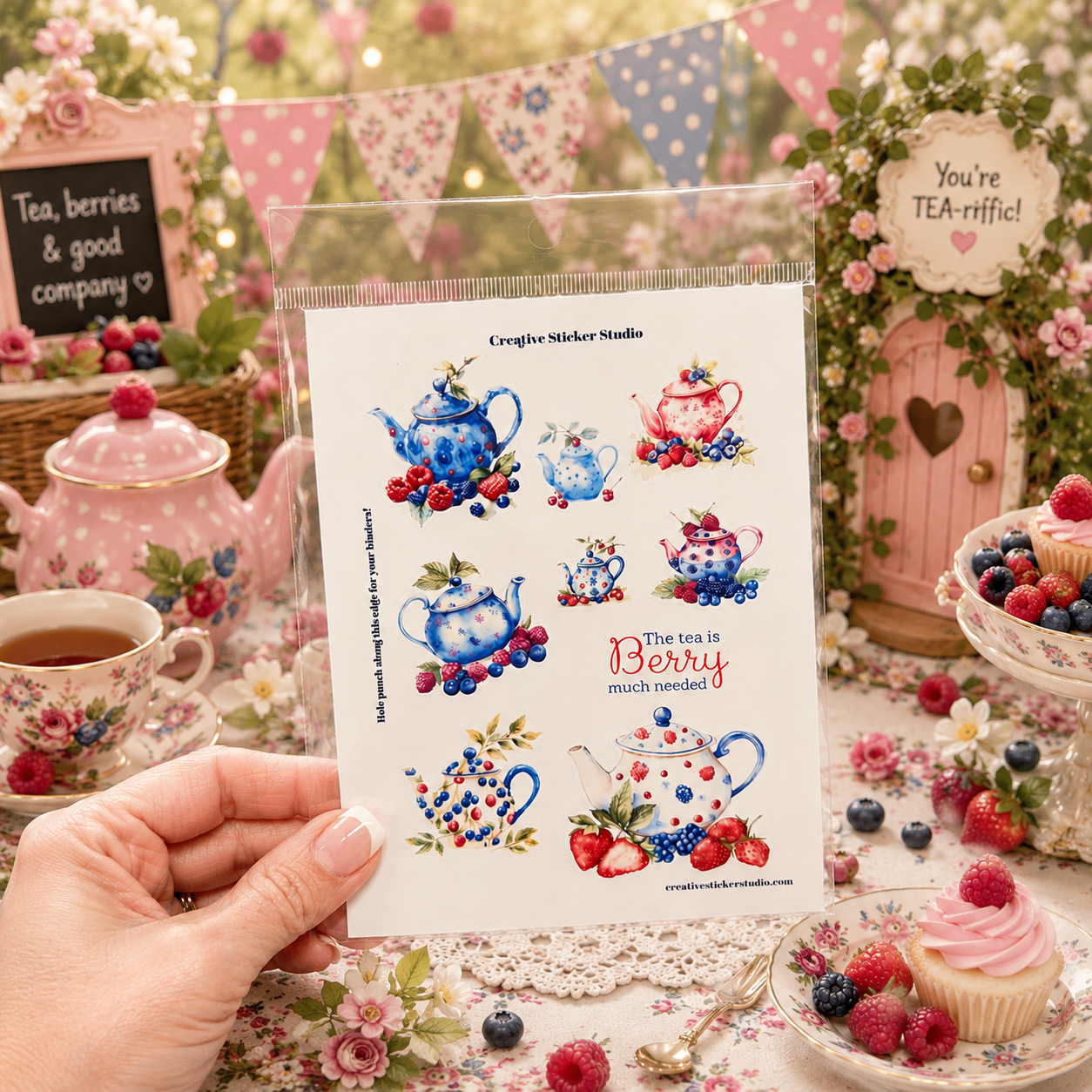 A hand holds a clear plastic sleeve containing a sheet of Creepy Cookey Studios tea-themed stickers, set against a highly styled, vintage-inspired tea party background with pink teapots, cupcakes, and floral decorations.