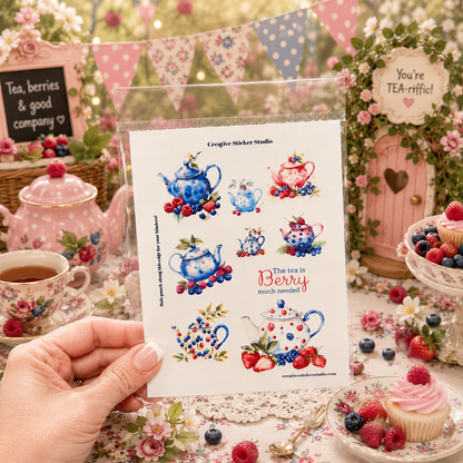 A hand holds a clear plastic sleeve containing a sheet of Creepy Cookey Studios tea-themed stickers, set against a highly styled, vintage-inspired tea party background with pink teapots, cupcakes, and floral decorations.