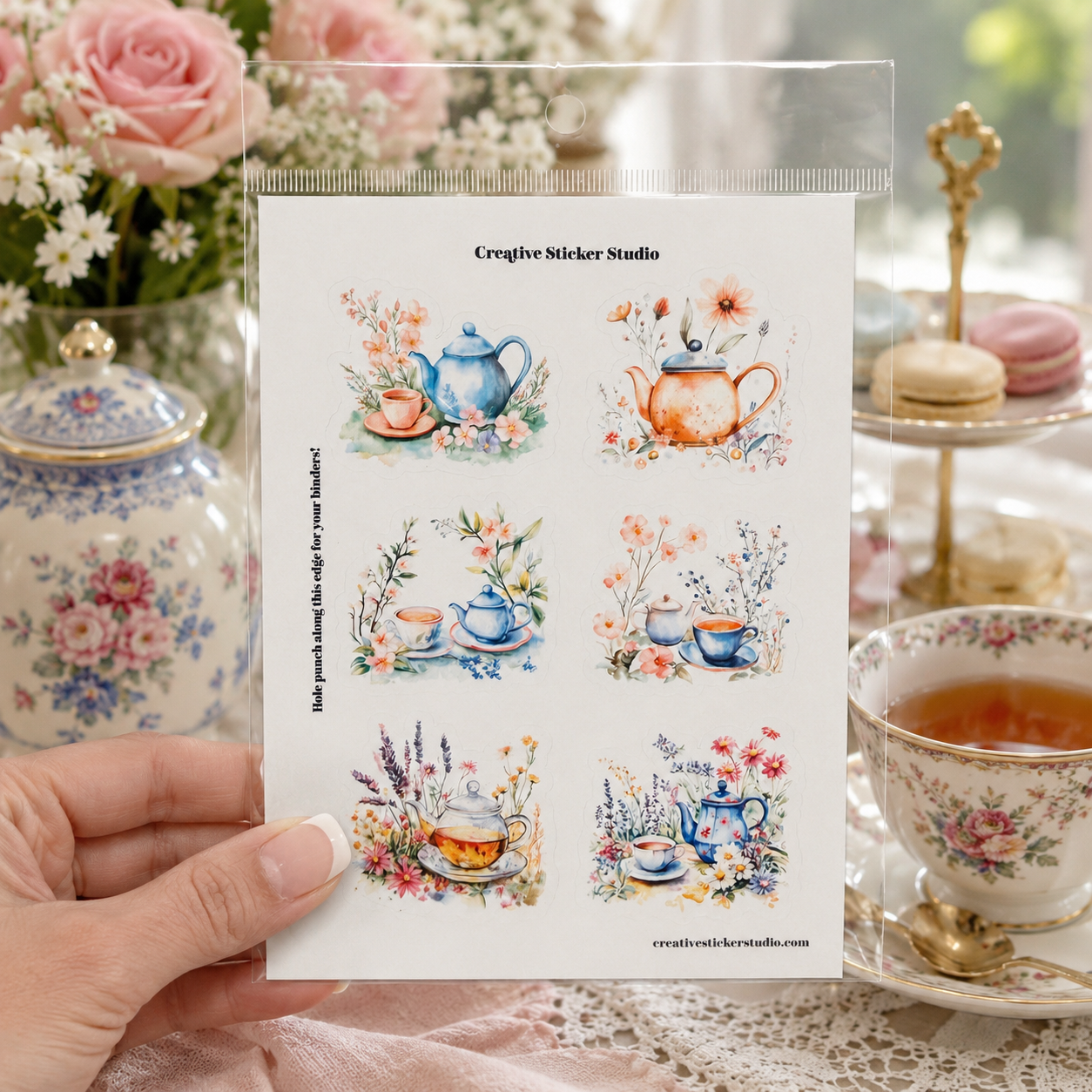 A hand holds a clear plastic sleeve containing a sheet of six watercolor tea-themed stickers. The stickers feature illustrations of teapots, cups, flowers, and tea sets in soft pastel colors.