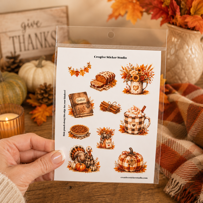 A hand holds a clear plastic sleeve containing a sheet of autumn-themed sticker designs. The sheet features illustrations of a loaf of bread, a mug of hot chocolate with whipped cream, a plaid pumpkin, a turkey, a small cabin, and fall garlands.